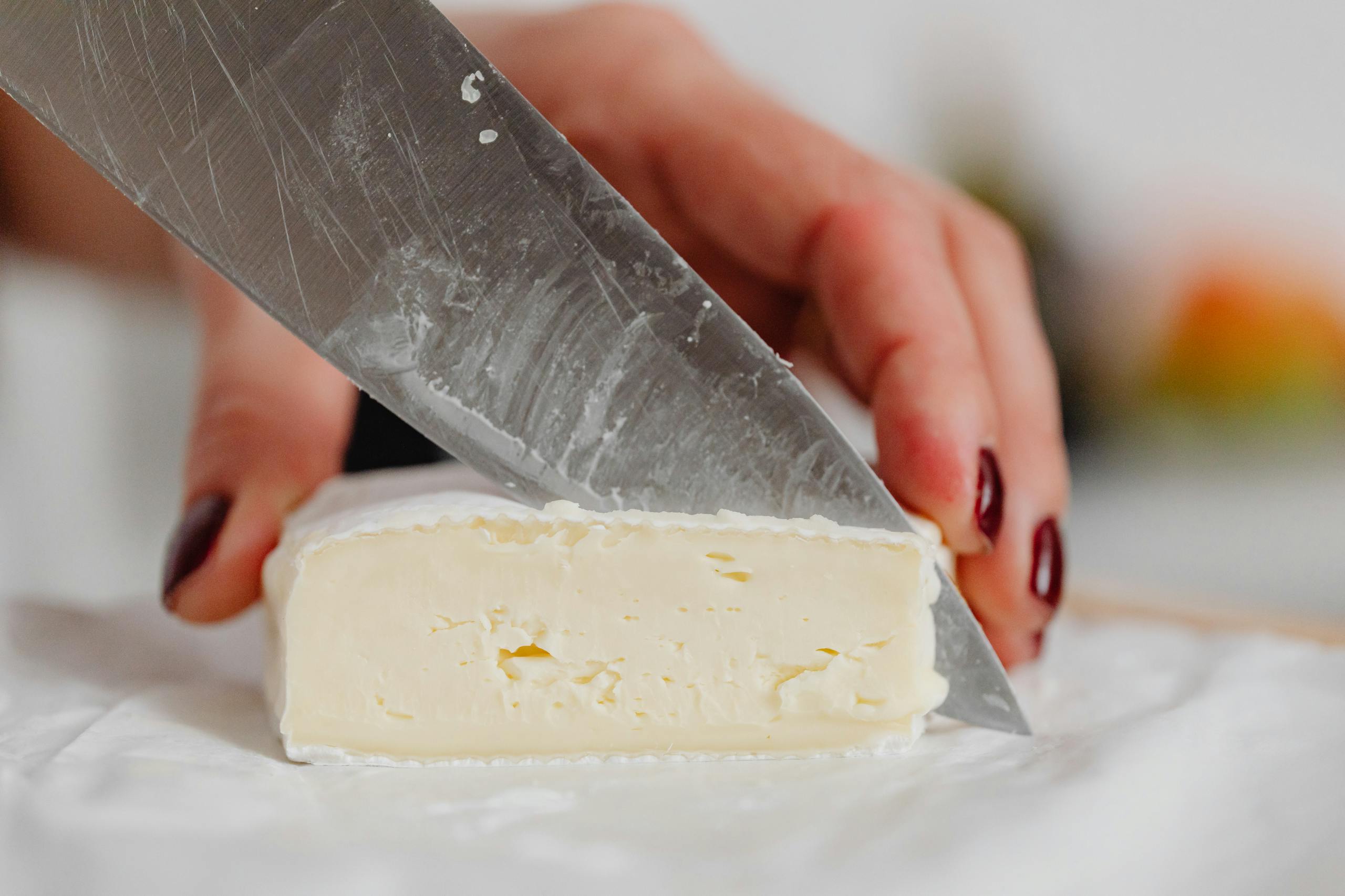 Close-up view of a hand slicing brie cheese with a knife on a white table, emphasizing creamy texture.